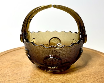 May include: A small, round, amber-colored glass basket with a scalloped rim and a textured exterior surface. The basket features a raised circular medallion design on the front and a hinged handle that arches over the top of the bowl.