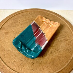 May include: A rectangular ceramic soap dish with a teal, orange, and red color scheme. The dish has a curved design on the ends and is placed on a round, brown, wooden surface. The dish is handmade and has a glossy finish.