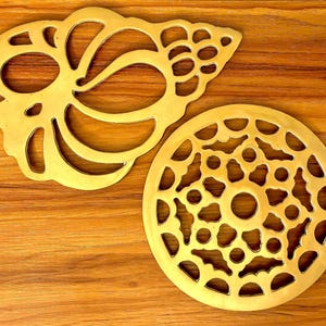 May include: Two decorative brass trivets with openwork designs. One is a stylized seashell shape, and the other is a circular shape with a central hole and leaf-like cutouts. The trivets are displayed on a wooden surface.