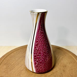 May include: A ceramic vase with a flared neck and a tapered body. The surface features a white base with vertical gold lines and a prominent, textured magenta panel with a crackled, bubble-like pattern. The rim is finished with a gold band.