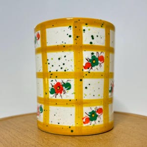 May include: A cylindrical ceramic container with a yellow grid pattern and floral accents. The grid is outlined in yellow, with white squares speckled with green and black dots. Red and green flowers are placed within the grid.