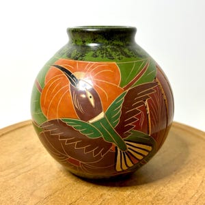 May include: A spherical ceramic vase featuring a hand-painted design of a hummingbird in flight. The bird is depicted in shades of brown, green, and yellow against a background of orange flowers and green leaves. The rim is a dark, mottled green.