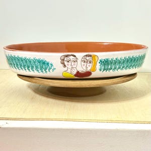 May include: A ceramic serving dish with a brown rim and a white interior. The exterior features a green brushstroke design and a painted illustration of two figures. The dish sits on a wooden base.