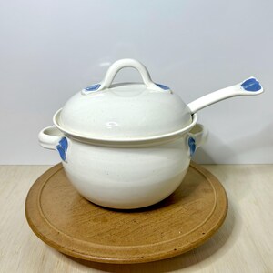 May include: A white ceramic soup tureen with a lid and a matching ladle. The tureen has blue floral accents and sits on a wooden serving tray. The ladle rests on the tureen's rim.