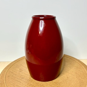 May include: A glossy, deep red vase with a rounded body and a narrow opening. The vase is set on a light brown, circular wooden surface. The vase's smooth surface reflects light, highlighting its shape and color.