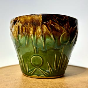 May include: A ceramic cup featuring a glossy glaze that transitions from a deep, dark brown at the rim to a vibrant forest green at the base. The surface is decorated with embossed circular shapes and pointed, grass-like vertical textures.