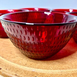 May include: A set of deep red glass bowls featuring a textured exterior with a repeating pattern of small, vertical indentations. The bowls have a smooth, rounded rim and a translucent quality that allows light to pass through the vibrant red glass.