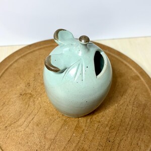 May include: A light blue ceramic container with a leaf design and a small opening. The container has a lid with a small knob and two ear-like handles. The handles and knob have a brown trim.