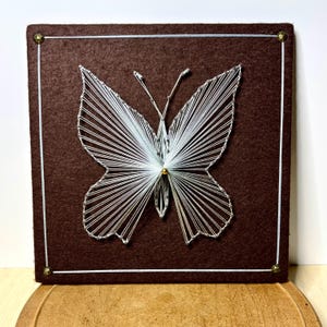 May include: A square brown felt board featuring a white string art butterfly design. The butterfly is created with thin white thread wrapped around small nails, forming a geometric pattern. A single gold bead sits at the center of the butterfly.