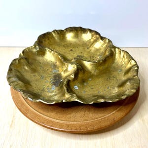 May include: A three-sectioned brass serving dish with a scalloped, shell-like edge and a weathered, tarnished gold finish. The dish features three distinct, rounded compartments joined together, resting on a circular, light brown wooden base.
