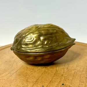 May include: A brass trinket box shaped like a walnut. The object features a textured, metallic gold surface with a realistic, irregular shell pattern and a horizontal seam running through the center, indicating a hinged lid design.