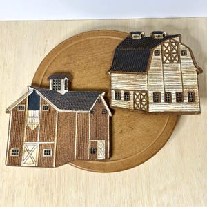 May include: Two ceramic barn-shaped decorative pieces with textured surfaces. One barn is brown with a dark roof and a cupola, while the other is cream-colored with a dark roof and detailed window and door patterns. Both items feature embossed lines.