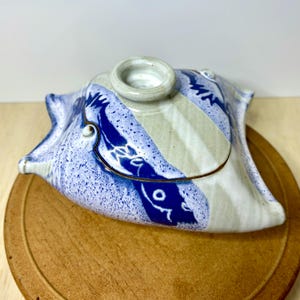 May include: A ceramic serving dish with a lid, featuring a unique, organic shape. The dish is primarily white with blue and tan stripes and speckled accents. A small, circular handle sits atop the lid. The dish rests on a round, wooden surface.