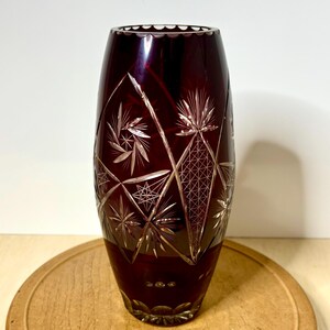 May include: A ruby red crystal vase with intricate etched designs. The vase features a scalloped rim and geometric patterns, including diamond shapes and starbursts. The vase is approximately 10 inches tall.