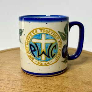 May include: Ceramic mug with a blue handle and rim, featuring the text "WE ARE WORKERS TOGETHER FOR CHRIST" around a cross and the letters "PWC" in a circle. The mug has a cream background with blue dots and green leaf and purple fruit designs.