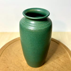 May include: A cylindrical ceramic vase with a matte forest green finish. The vase features a slightly flared rim and two subtle, horizontal incised rings encircling the neck area, providing a simple texture to the smooth, solid-colored surface.