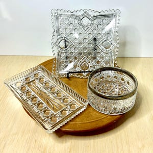 May include: A collection of vintage clear glass serving pieces with intricate cut patterns. Includes a square tray, a rectangular tray, and a round dish with a silver rim. The pieces are arranged on a wooden surface.