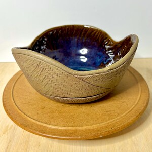 May include: A ceramic bowl with a textured, light brown exterior featuring a herringbone pattern and a smooth, glossy interior glazed in deep shades of blue and purple. The rim of the bowl has an irregular, wavy shape with a thick, rounded edge.