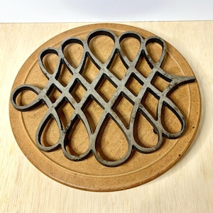 May include: A vintage metal trivet with a decorative openwork design in a teardrop and diamond pattern. The trivet is a dark gray color and rests on a round, light brown ceramic base. The trivet is designed to protect surfaces from hot cookware.