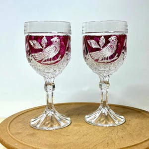 May include: Two clear crystal wine glasses with a ruby red interior. Each glass features a detailed etched design of a bird perched on a branch with leaves. The glasses are displayed on a wooden surface.