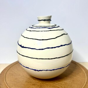 May include: A spherical ceramic vase with a smooth off-white finish, featuring a series of horizontal, raised dark blue lines that wrap around the body. The vase has a small, narrow neck opening at the top and a rounded, bulbous base shape.