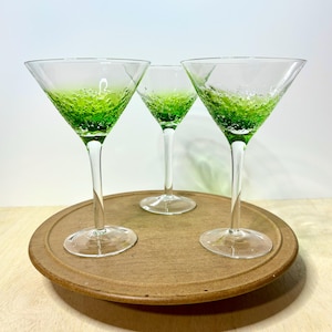 May include: Three martini glasses with clear stems and bases, featuring a wide, conical bowl. The lower portion of each bowl is crafted from textured green glass with visible air bubbles, transitioning into a smooth, clear glass rim.