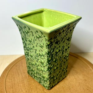 May include: A square-shaped, green ceramic vase with a textured surface. The vase has a flared design and a bright green interior. The exterior features a mottled pattern of darker and lighter green hues, creating a unique, organic look. The vase is sitting on a wooden surface.
