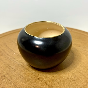 May include: A small, round, black ceramic bowl with a gold-colored rim. The interior of the bowl is a light beige color. The bowl is sitting on a wooden surface.