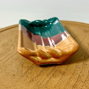 May include: A small, ceramic soap dish with a rectangular shape and a twisted rope design around the edges. The dish is painted with horizontal stripes of green, burgundy, and tan colors.