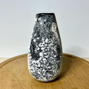 May include: A black and white speckled ceramic vase with a narrow neck. The vase has a textured surface and a dark rim. The vase is displayed on a wooden surface.