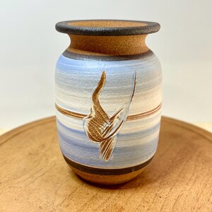 May include: A ceramic vase with a textured brown rim and base, featuring horizontal bands of light blue, white, and tan. The center of the vase displays a carved, abstract brown design that resembles a stylized bird or wing shape in relief.