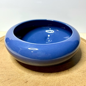 May include: A round, low-profile ceramic bowl with a smooth, glossy finish in a deep, vibrant blue color. The bowl features a wide, curved rim that slopes inward toward a deep, circular basin, creating a simple, open, and functional design.