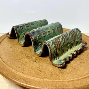 May include: Three handmade ceramic soap dishes with a wavy design. The dishes are glazed in shades of green and brown, with a textured surface. They are arranged on a round, wooden tray.