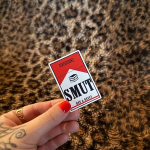 May include: A rectangular sticker with a red and white design, featuring the word "SMUT" in bold black letters, and the words "HOT & HEAVY" below. The sticker also has the word "UNFILTERED" in a red box.