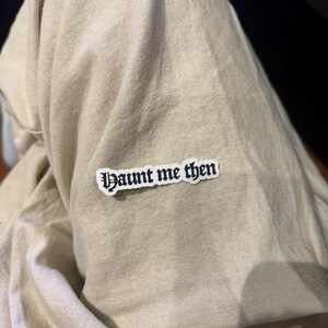 May include: A white sticker with the text "Haunt me then" in a gothic font is affixed to a beige fabric. The sticker has a black border and the text is in black. The fabric appears to be a linen or cotton blend.