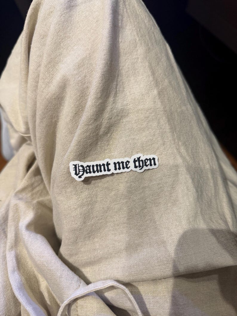 May include: A white sticker with the text "Haunt me then" in a gothic font is affixed to a beige fabric. The sticker has a black border and the text is in black. The fabric appears to be a linen or cotton blend.