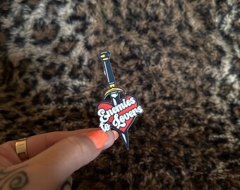 May include: A red heart with a black dagger piercing through it. The heart has the text "Enemies to Lovers" in a yellow and white font.