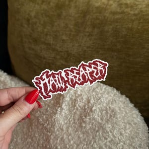 May include: A red and white sticker with the word "TAILORED" in a stylized, gothic font. The sticker is held by a hand with red nail polish, against a backdrop of a white, fluffy surface and a gold-colored pillow.