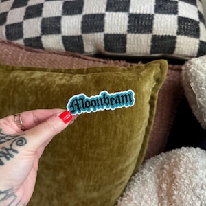 May include: A sticker with the word "Moonbeam" in a blue and white font. The sticker is held in front of a green velvet pillow, with a black and white checkered pillow in the background.
