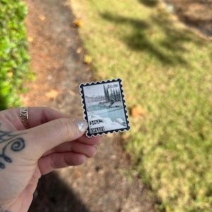 May include: A black and white sticker with a vintage postage stamp design. The sticker features a scenic landscape with trees and a body of water. The text "Still Not Home" is printed on the sticker.