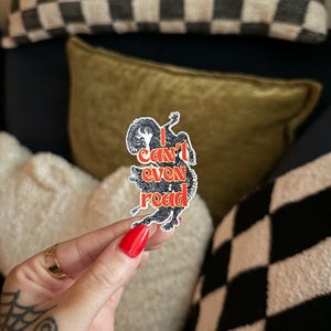 May include: A sticker with the text "I can't even read" in red and black, featuring a stylized illustration of a wolf-like creature. The sticker is held against a backdrop of pillows with checkered and solid patterns.