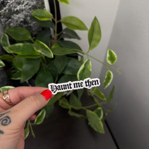 May include: A white sticker with the text "Haunt me then" in a gothic font is held in front of a green plant with white-edged leaves. The sticker has a black border. The person holding the sticker has red nail polish.