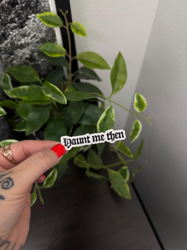 May include: A white sticker with the text "Haunt me then" in a gothic font is held in front of a green plant with white-edged leaves. The sticker has a black border. The person holding the sticker has red nail polish.
