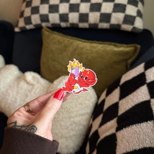 May include: A red devil sticker with a yellow belly button and a purple book with flames. The sticker is held in front of a background with pillows in black and white checkerboard, olive green, and white.