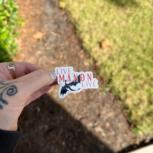 May include: A white sticker with a red and black design. The sticker features a black silhouette of a dragon with the text "LIVE MANON LIVE" in red.