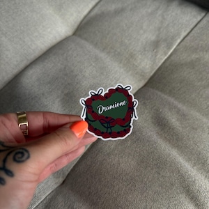 May include: A green heart-shaped sticker with red and black details. The sticker has the word "Dramione" written on it.