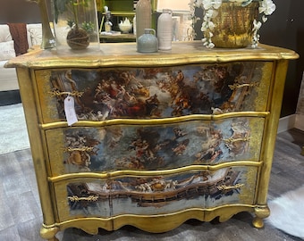May include: A gold painted chest of drawers with three drawers. Each drawer has a decorative design of a scene with angels and clouds. The chest of drawers has a curved front and sits on four legs.