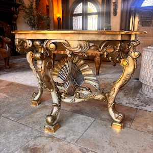 May include: Ornate, antique side table with a gold and cream finish. The table features elaborate carvings, including a shell-shaped design on the front and scrolled legs. The tabletop is rectangular. A luxurious piece of furniture.