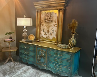 May include: A teal blue dresser with gold accents and ornate carvings. The dresser has six drawers and a center cabinet with a carved cherub design. The dresser is sitting on a white shag rug in front of a gold-framed mirror with a cherub design.