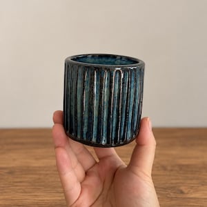 May include: A cylindrical ceramic cup featuring a deep blue and black mottled glaze with a vertical ribbed texture. The exterior surface displays a glossy finish with dark brown undertones visible within the grooves of the vertical ridges.